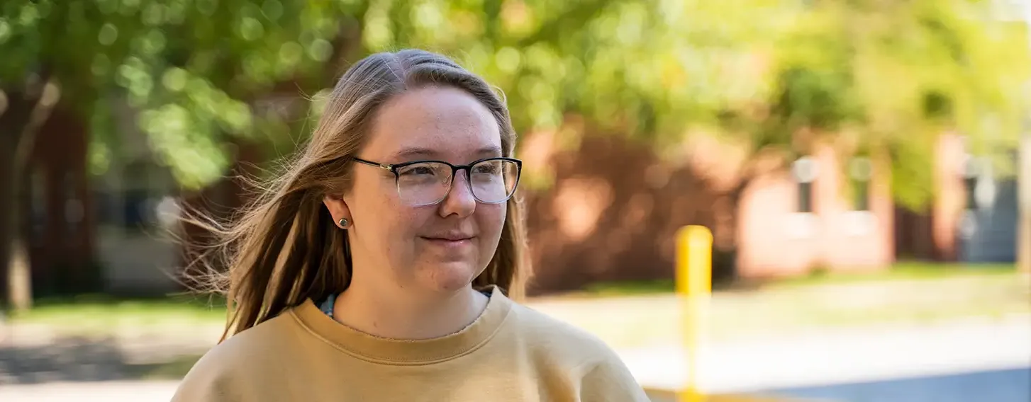 A girl with glasses and a gold sweatshirt walking outside on a sunny day.